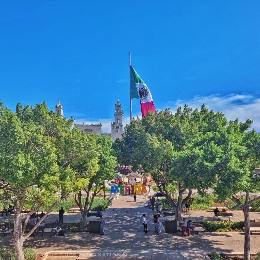 view over Mérida's plaza - Mexican flag flying, cathedral in distance, Merida letters visible through trees