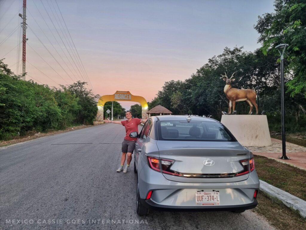 person in red tshirt and grey shorts leaning on a silver car on an empty road at dusk