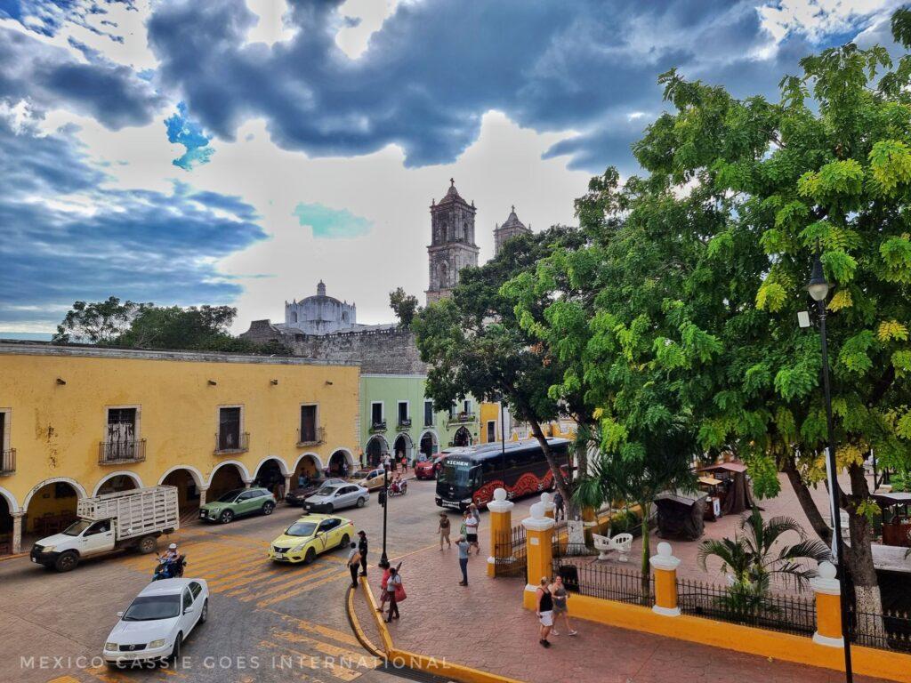 looking down over a road with parked cars and 2 cars driving around the corner of a plaza. big tree on right covering most of a cathedral