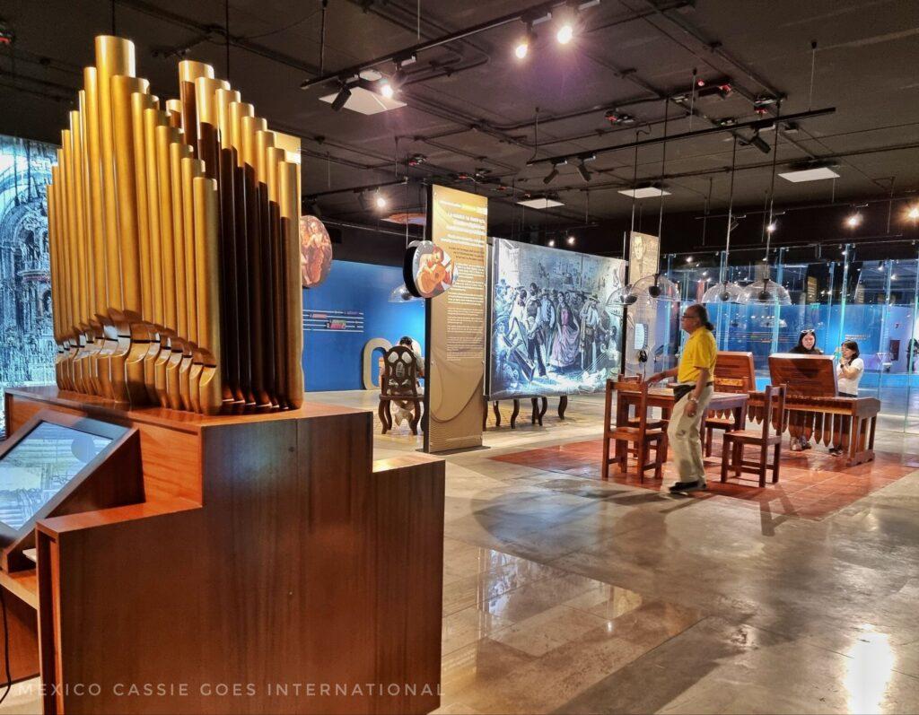 view of the inside of the music museum. organ on left, table with 4 chairs, man in yellow shirt. 2 girls at another table looking at exhibition