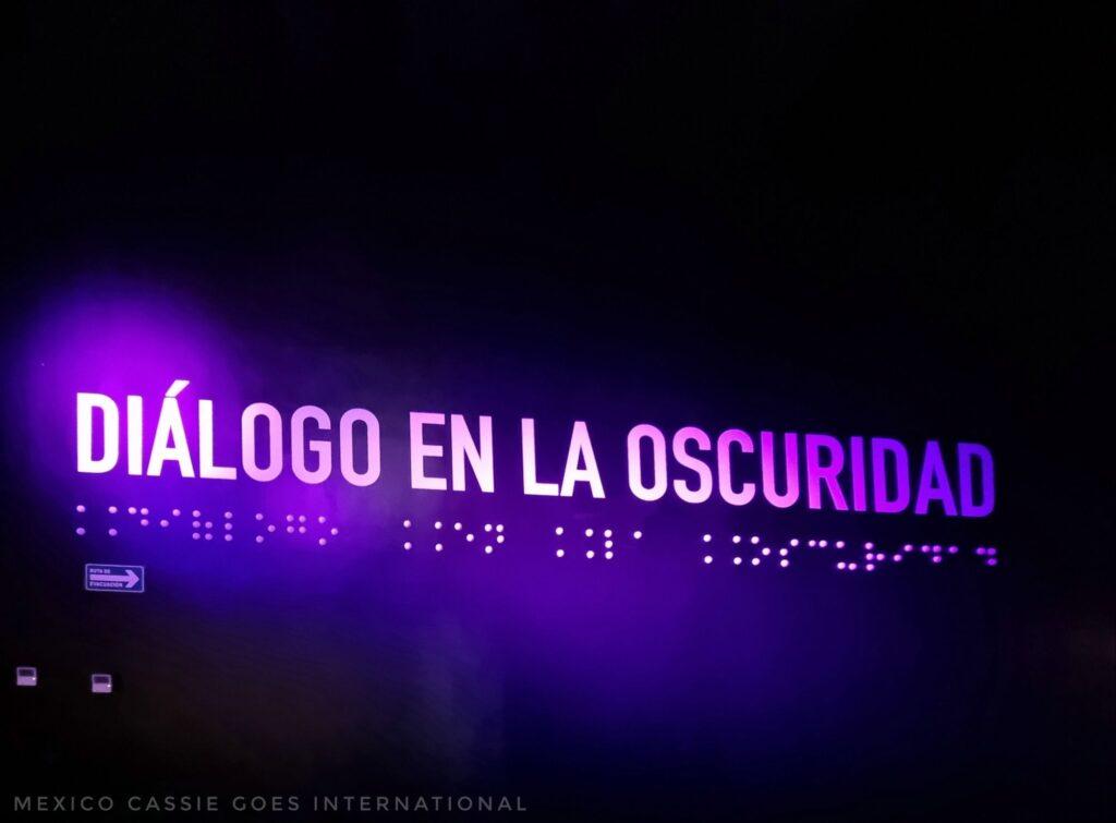 Words "Diálogo en la Oscuridad (braille underneath) in luminous purple with glow into the black background