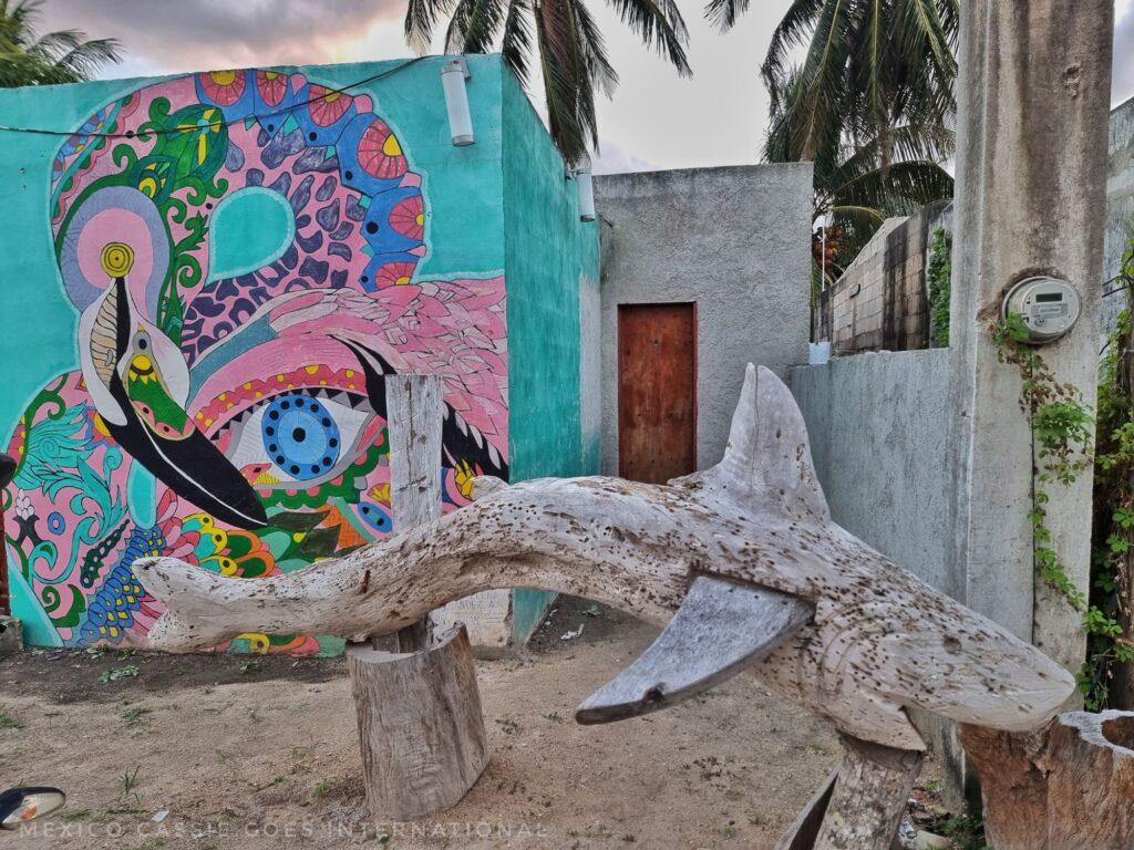 green wall with multicoloured flamingo painted on it, wooden shark sculpture outside