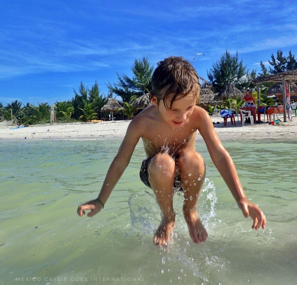 small child jumping out of green sea, blue sky, white sand and green tres behind