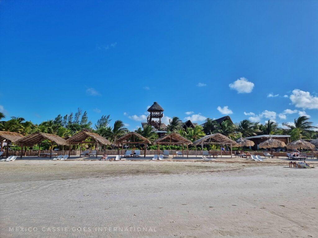 shot taken while standing in shallow sea looking back at beach with beach huts (thatched), green trees and blue sky