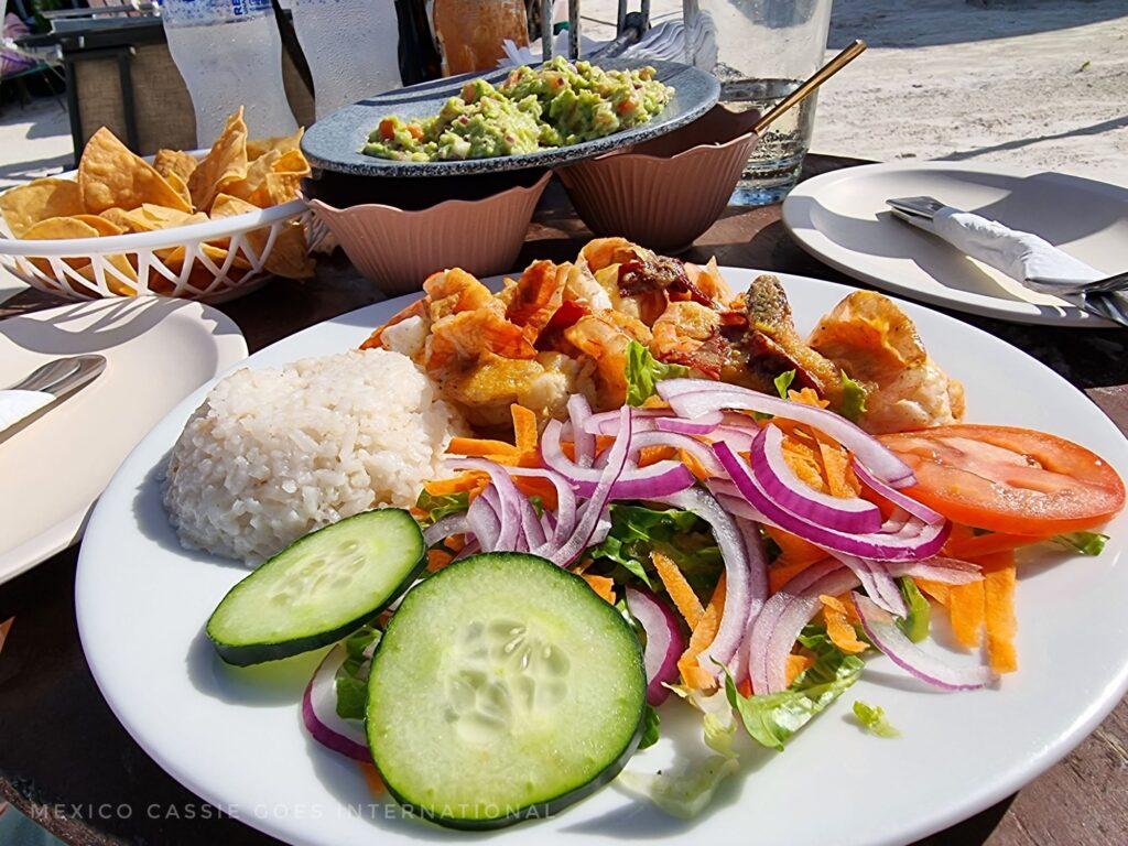plate of prawns with rice, cucumber, tomato, onion, guacamole in bowl behind