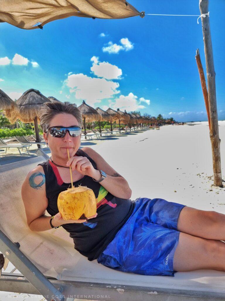 Cassie drinking coconut water from a coconut while sitting on a lounger under a shade on the beach