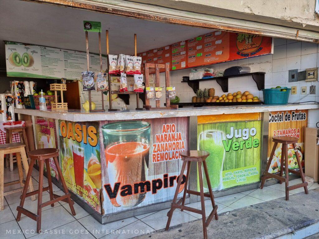 typical mexican juice bar, stools around bar - signs for vampiro, jugo verde, oasis, big kiwi picture on back wall, pile of oranges and pineapples on side