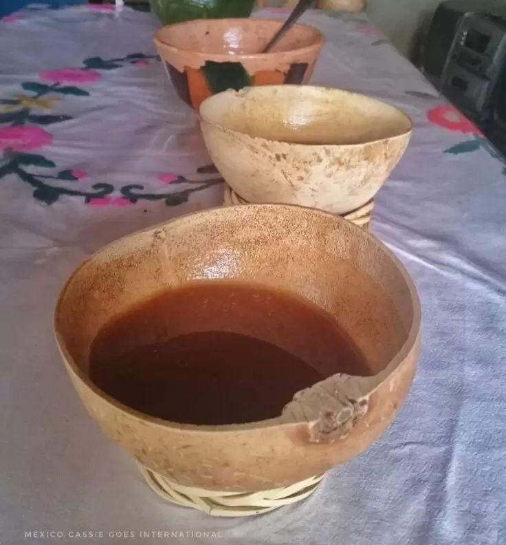 2 gourds with a brown liquid on a table with white tablecloth, regular cup there too