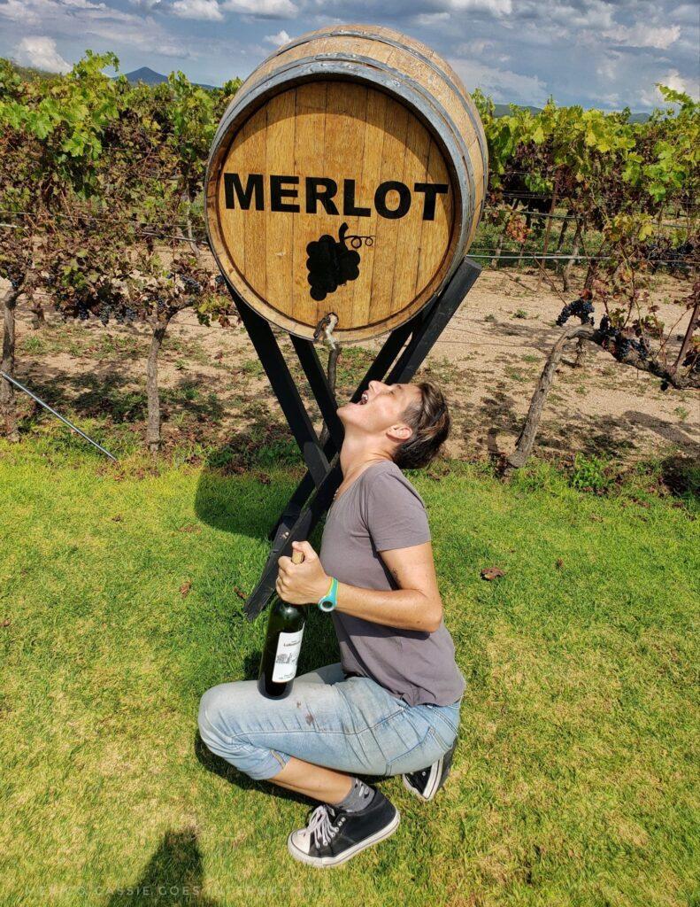 Cassie under a wine cask that says "Merlot" on it, mouth open, bottle of wine in hand