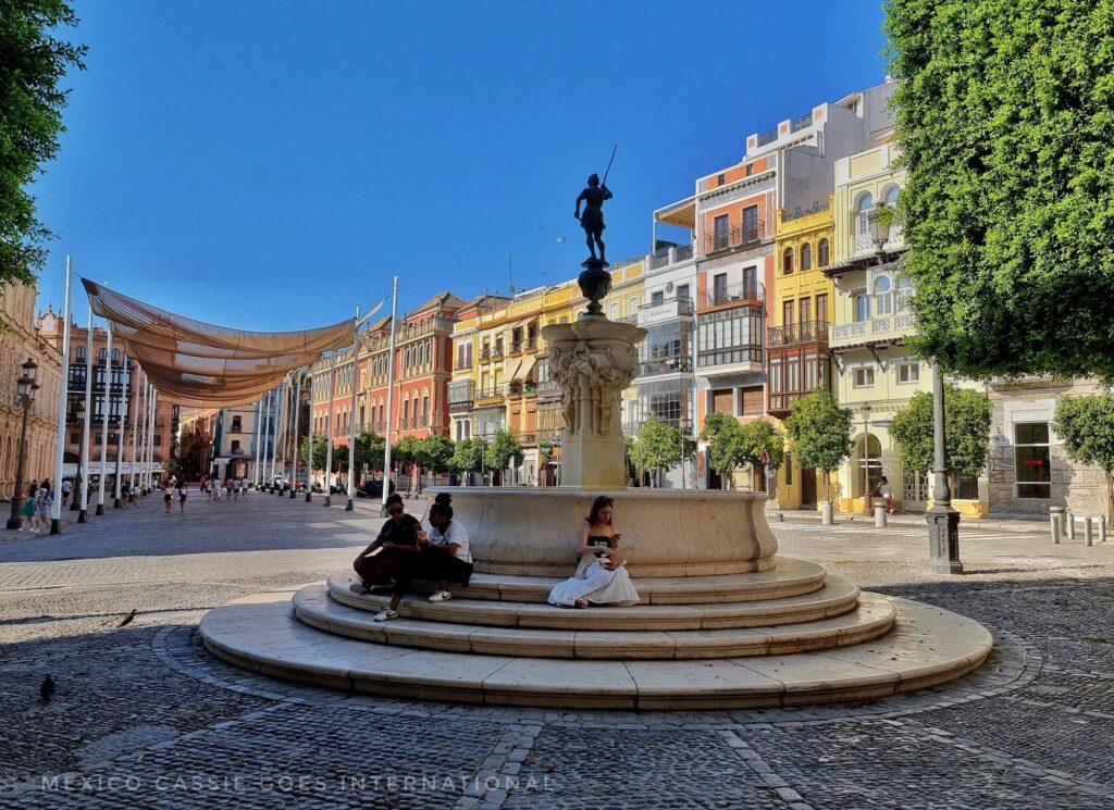 almost empty plaza, small fountain with people sitting around it. houses on side