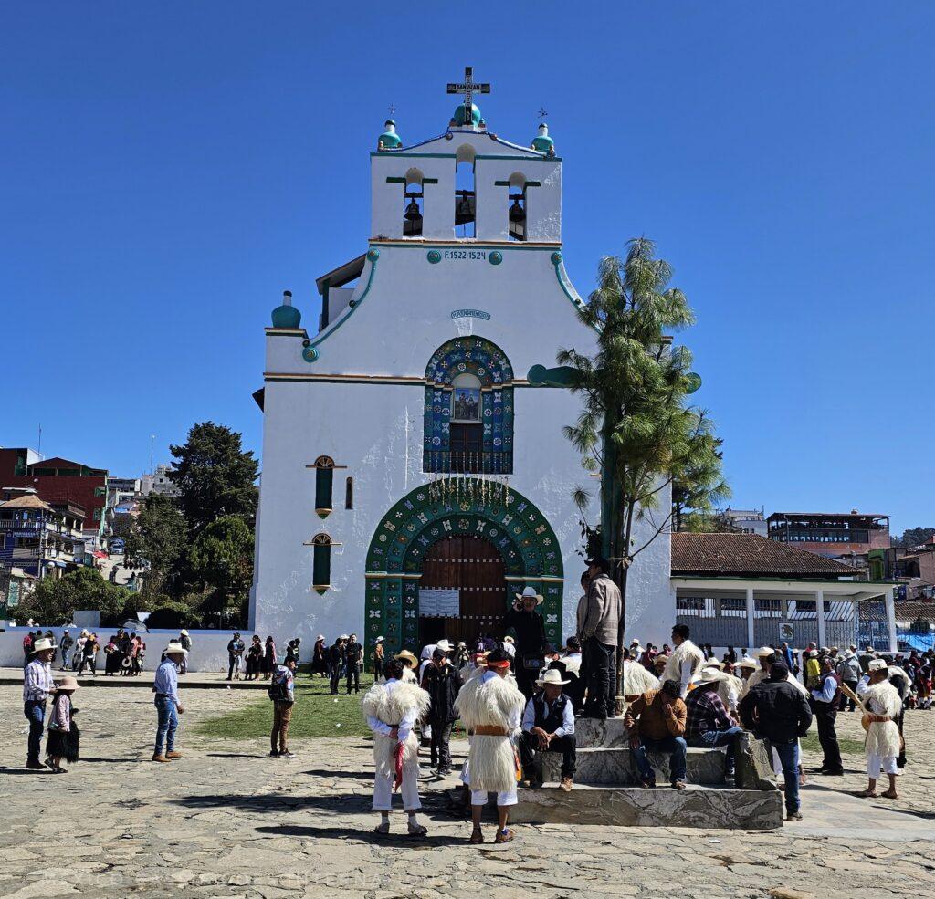 white church with green 'trim' - people milling around in plaza in front in white traditional costumes
