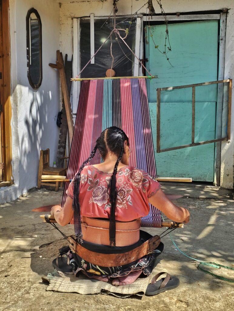 back of woman in traditional clothing with two long plaits. she's sitting on the ground weaving