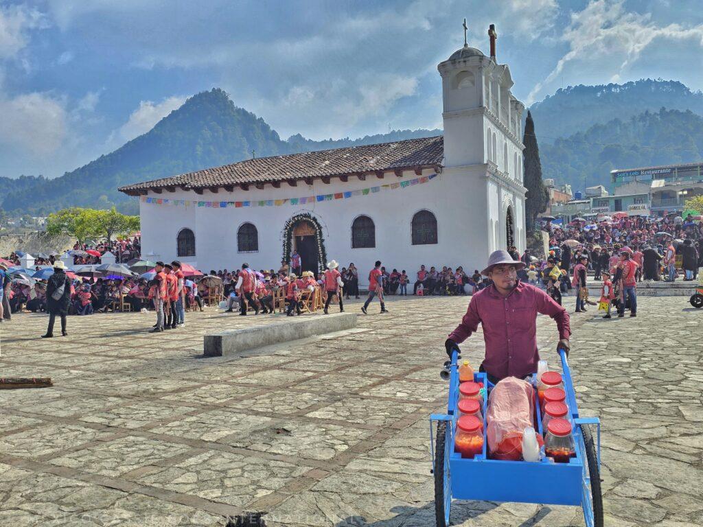 large plaza, people around church in background, foreground is a guy pushing a blue cart with drinks