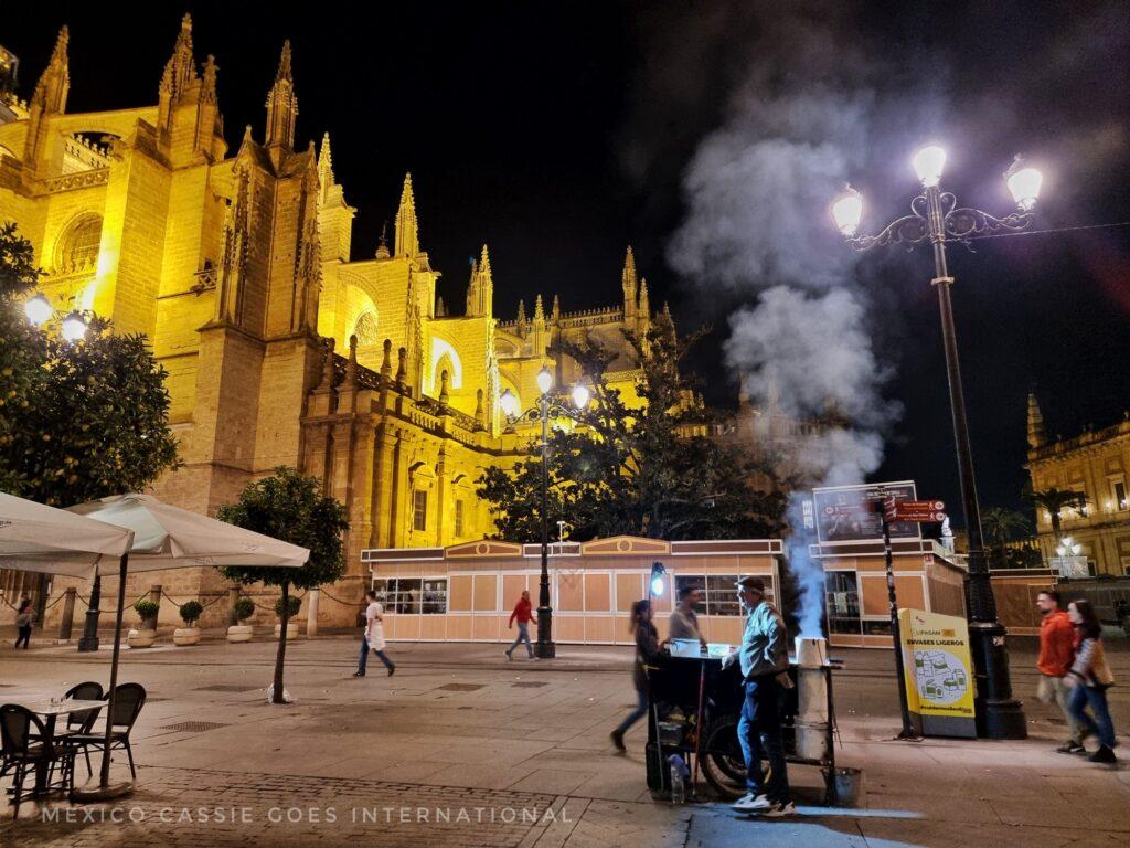 Seville cathedral lit up in the dark - chestnut stand in front with smoke rising from it