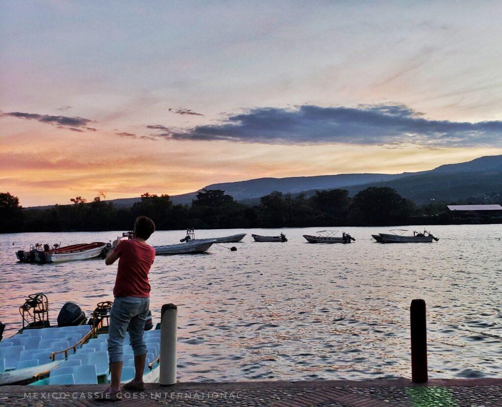 Cassie standing on bank of river taking a photo of sunset. small boats on river