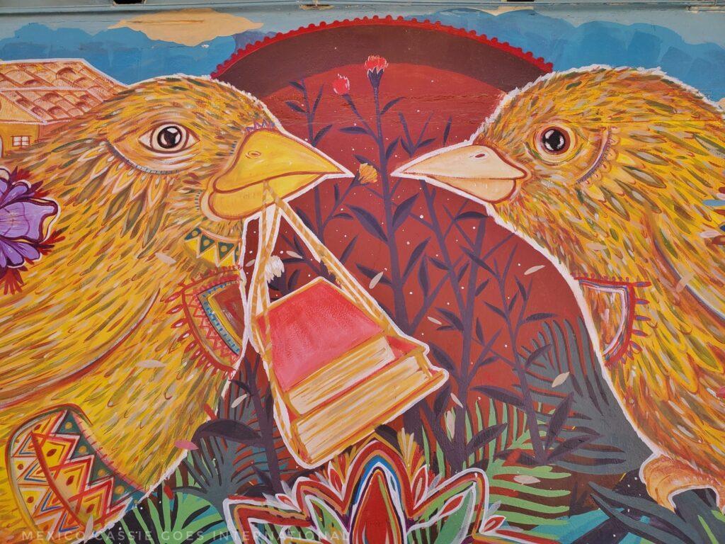 street art - two birds looking at each other, 1 caring 2 books in a strap from its beak