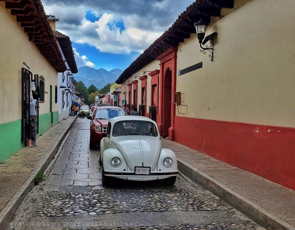 white VW and red car behind it on cobbled street- buildings on either side and mountains in distance
