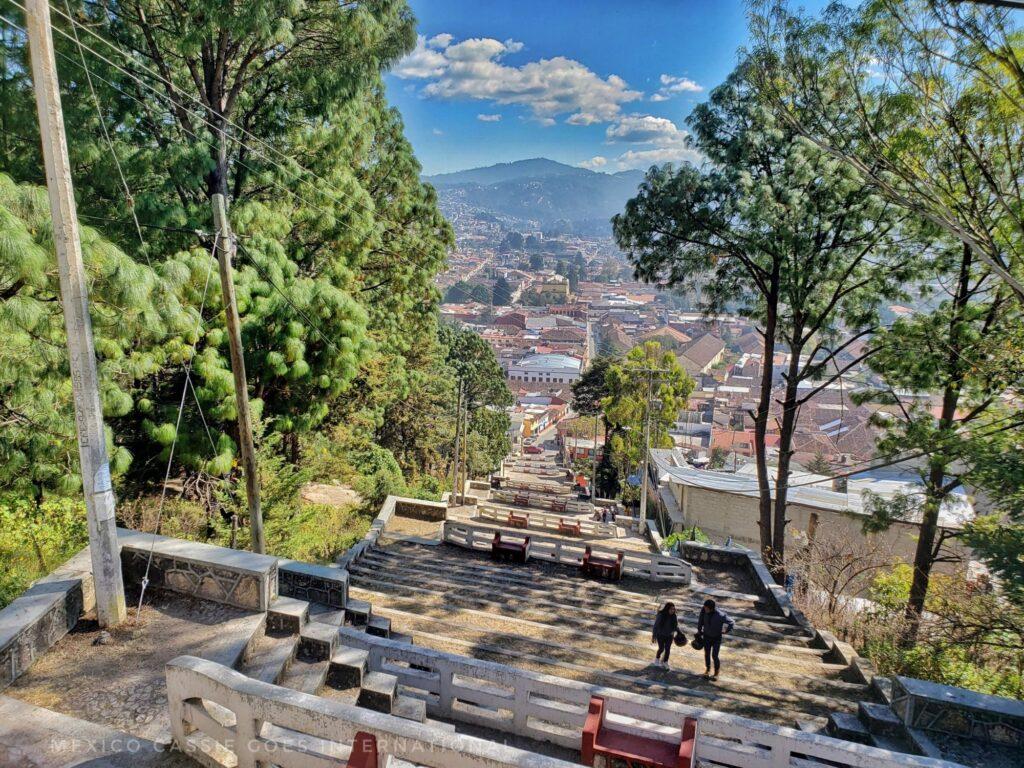 looking down a massively long flight of outdoor steps with trees on either side & mountains in distance behind town