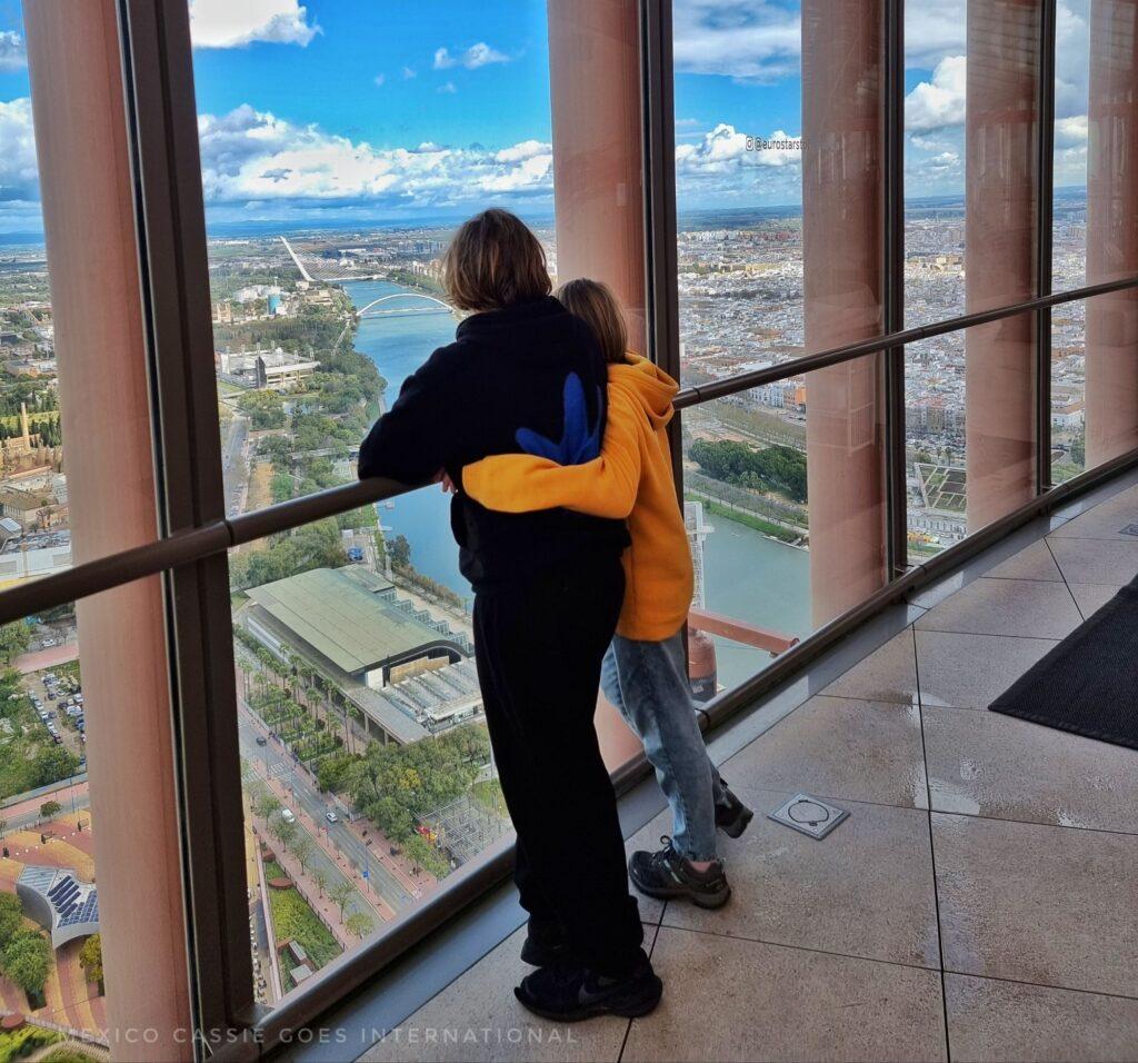2 kids standing together facing away from camera looking out over city and river 