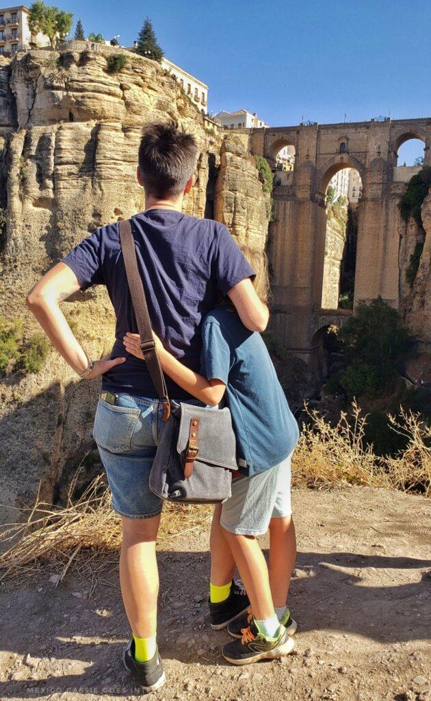 adult in blue tshirt and denim shorts and kid in shorts facing away from camera. both looking at the enormous puente nuevo in ronda