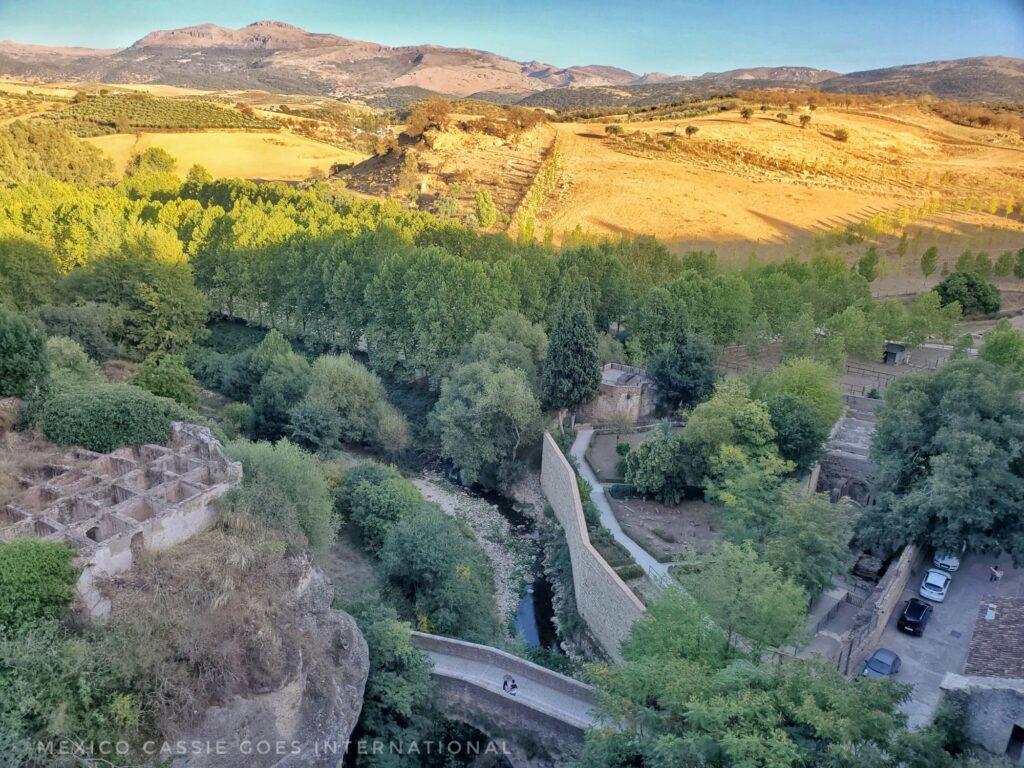 looking down over small bridge and stream - trees in shadow but fields behind still in sun. row of cars by Arab baths