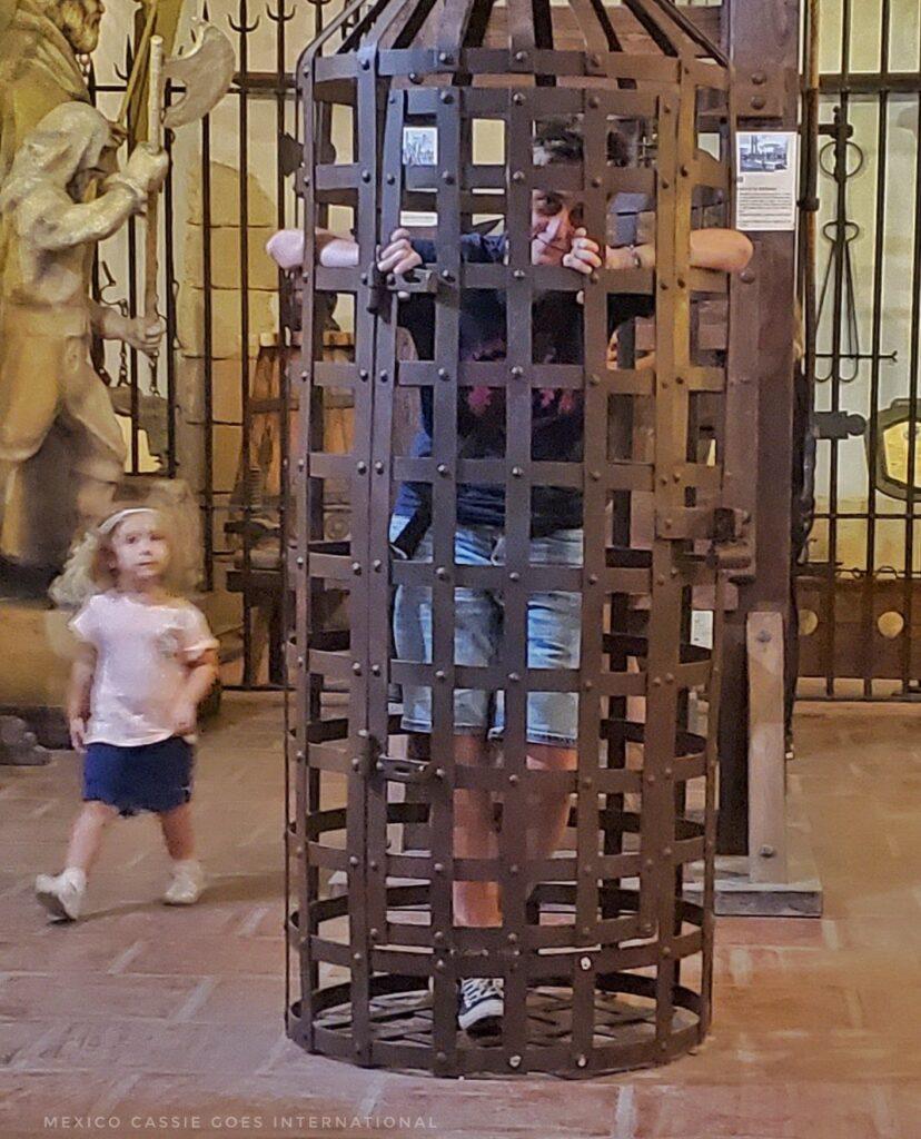 cassie locked in a medieval torture device cell , tiny child on one side walking by