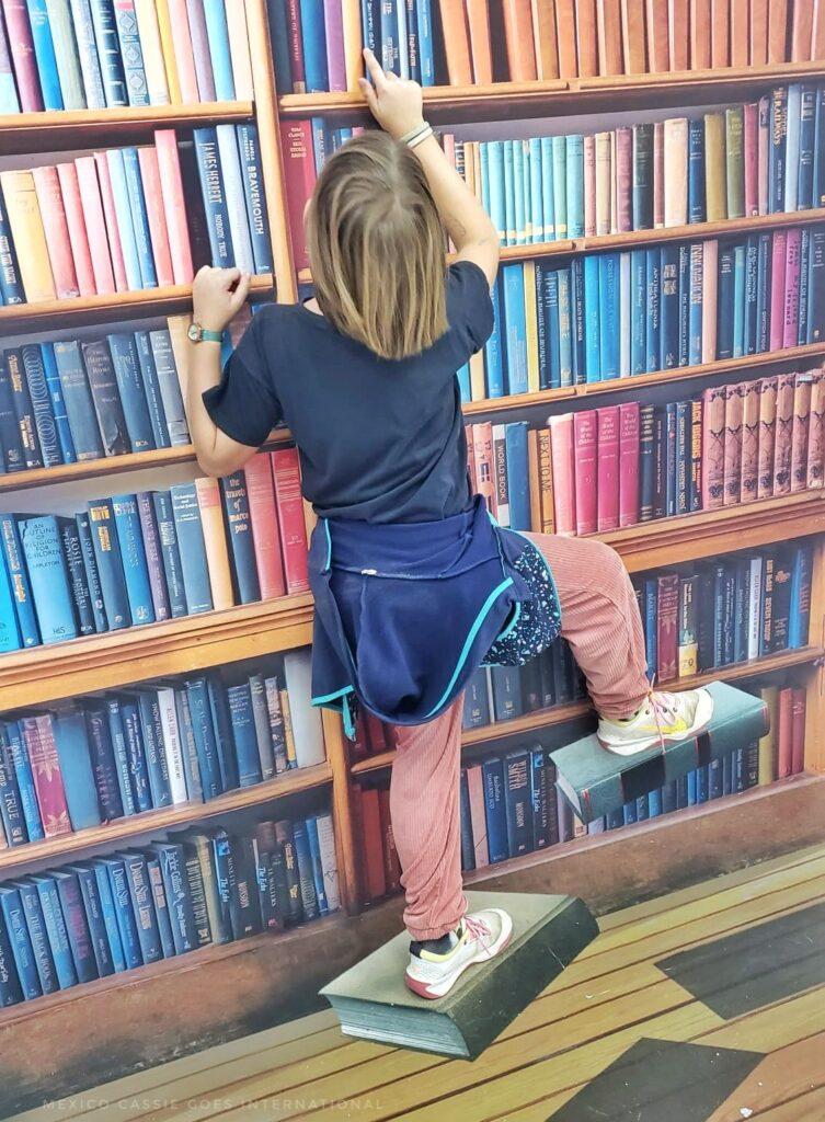 kid climbing on books (it's an illusion) to find a book on the shelf