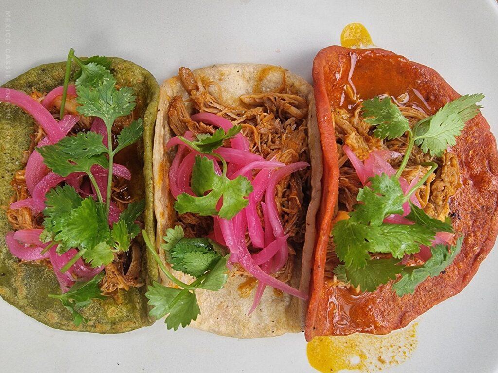 plate of 3 tacos - tortillas are black, white, red. all with meat, red onion and cilantro on them