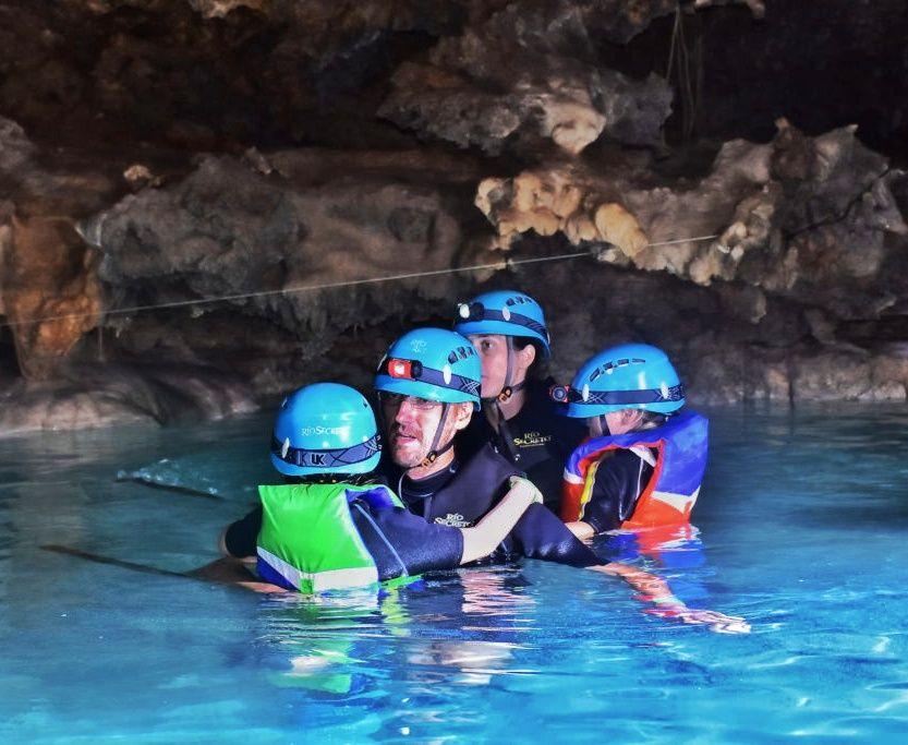 family of 2 adults and 2 kids in clear blue water in a cave -all in life jackets and blue helmets