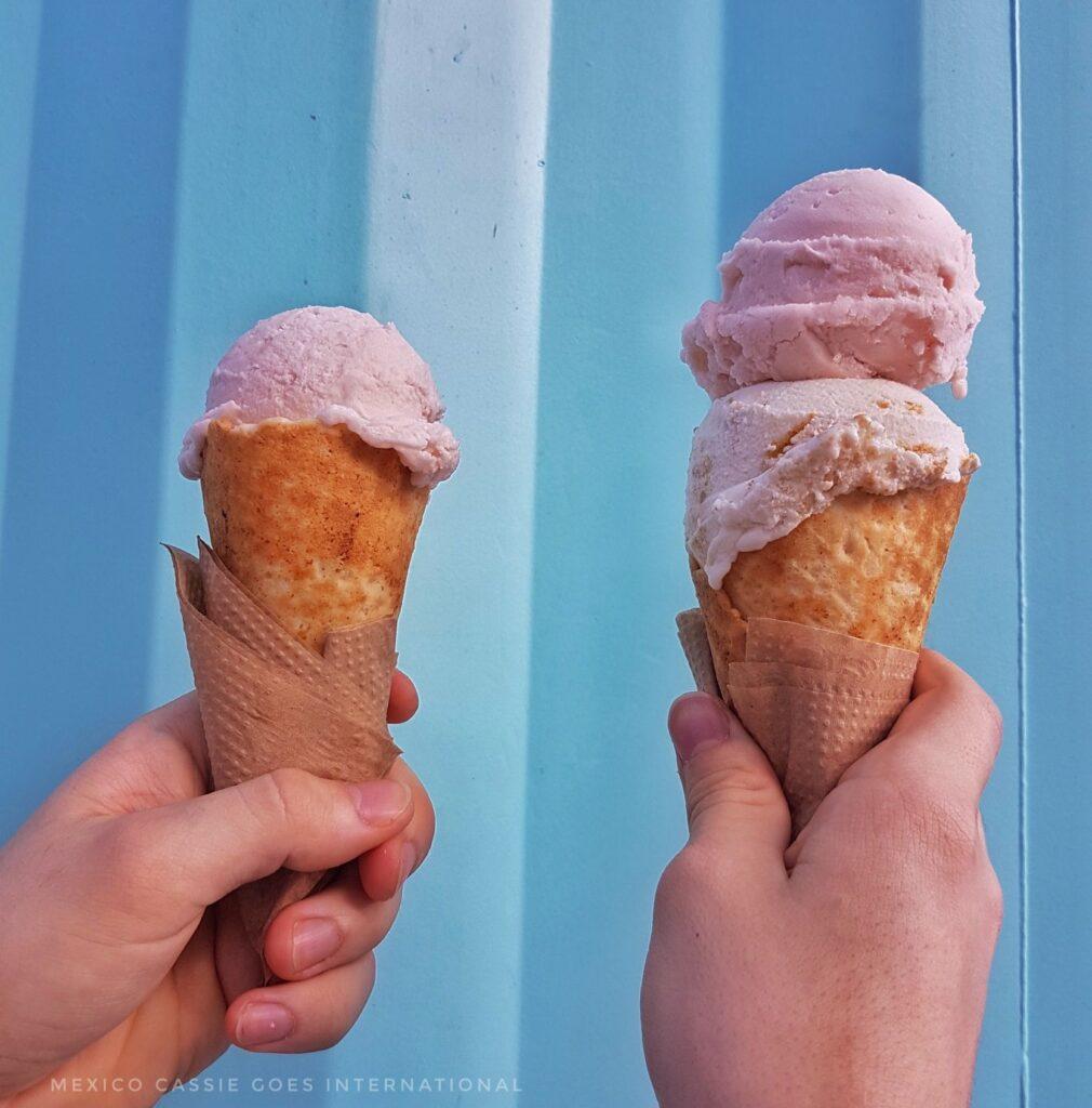 2 hands holding up ice cream cones (1 with 1 scoop, 1 with 2) against a blue striped wall