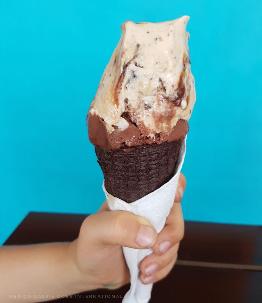 ice cream in a chocolate cone wrapped in a white serviette held by a hand against a turquoise  wall
