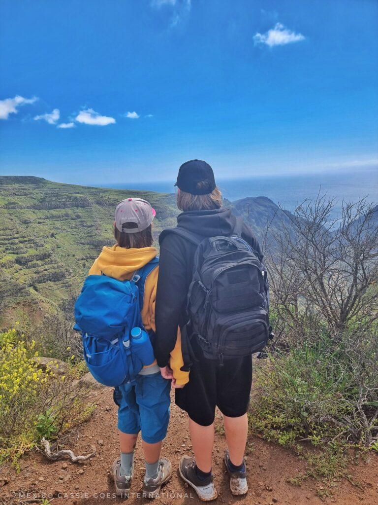2 kids in hoodies, caps and rucksacks looking out of view over a valley