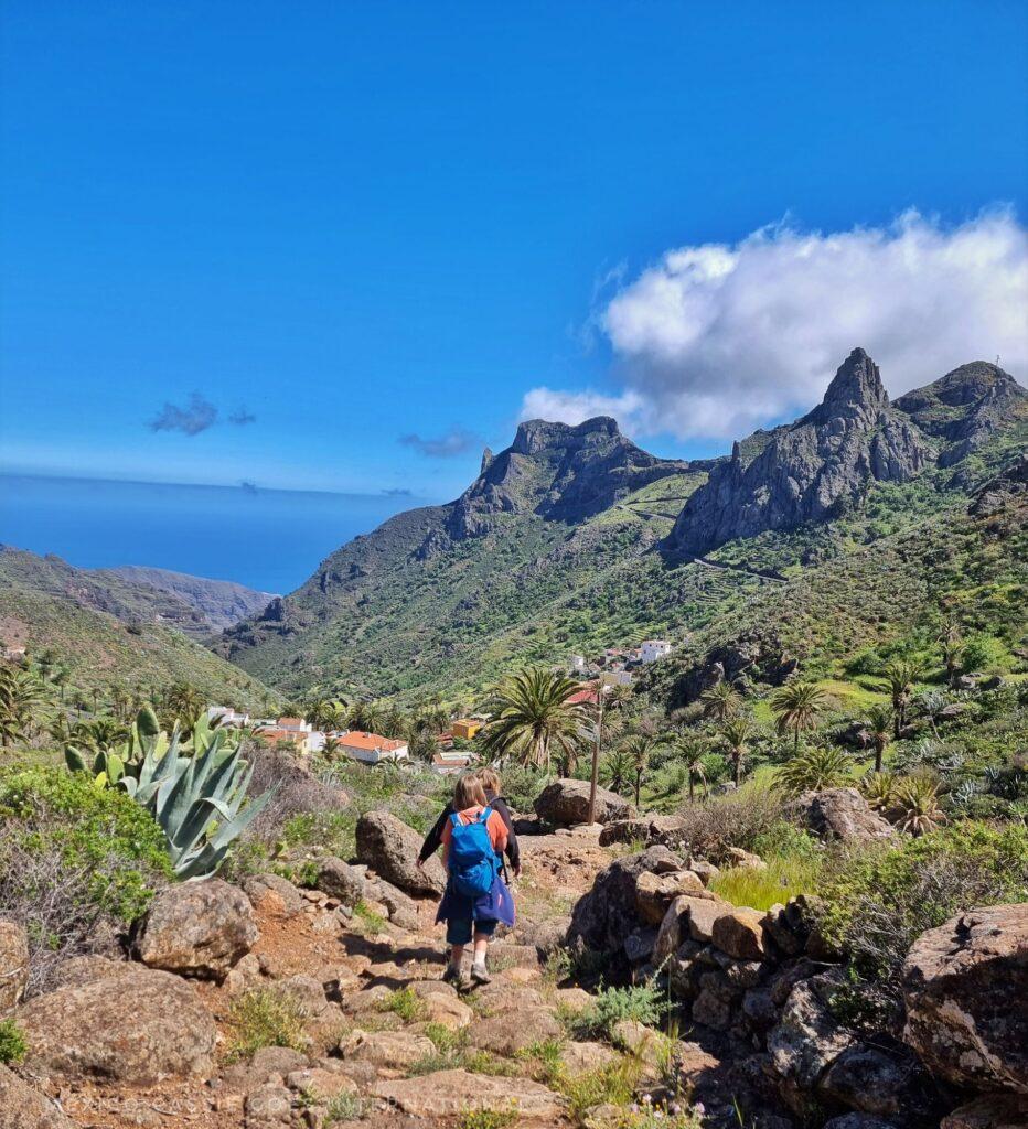 2 kids walking down a path (away from the camera), mountains around, sea in distance, cloud over mountain, houses in near distance, palm trees