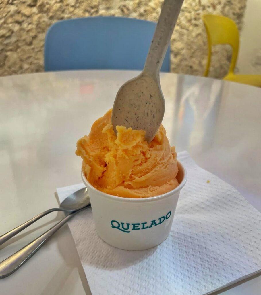 orange colour ice cream in a white cup that has "Quelado" written on it in blue. Wooden spoon in the ice cream, metal spoon and napkin on table