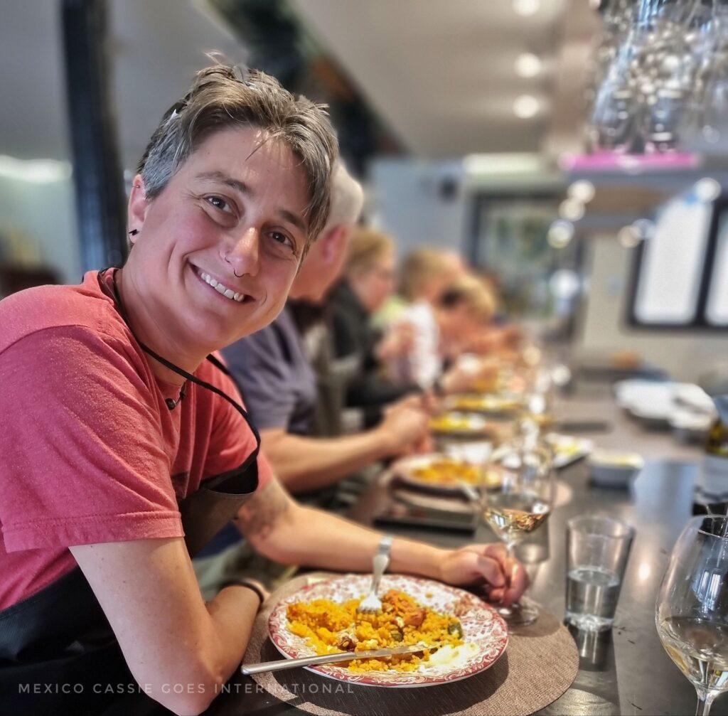cassie in a red tshirt eating paella (and looking directly at the camera). people in a seated line beside her all blurred