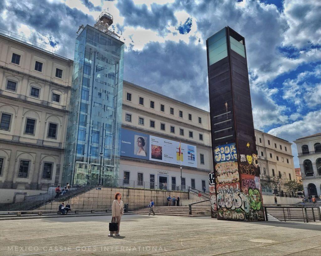 outside of the Reina Sofia museum in Madrid - glass lift space on outside of building, also a square iron tower covered in graffiti on the plaza- woman walking across plaza