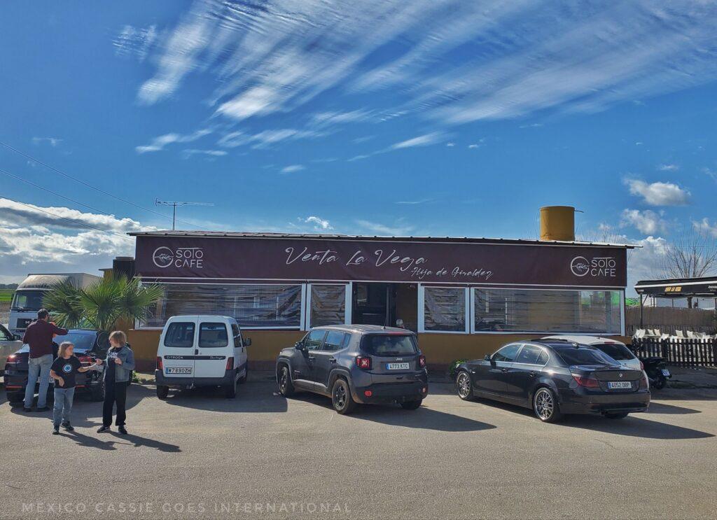 a one storey building with 4 cars parked outside it. 3 people in view. (building is a venta - a restaurant by the side of the road)