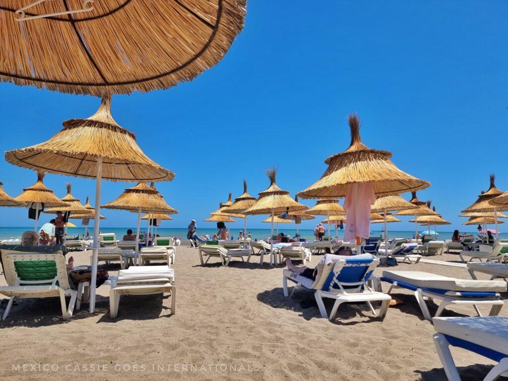beach scene, blue sky, sandy beach with white loungers and straw umbrella shades