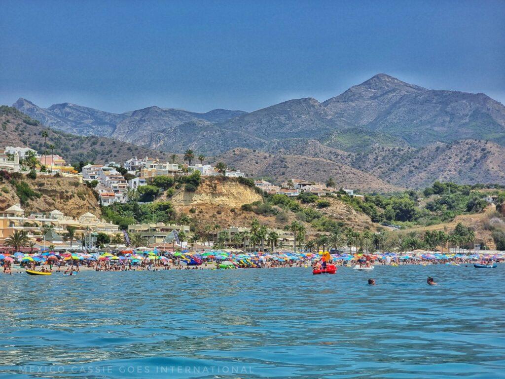 very clear blue water (photo taken from a paddle board looking back at Malaga coastline), mountains, beach with lots of umbrellas on it, red kayak
