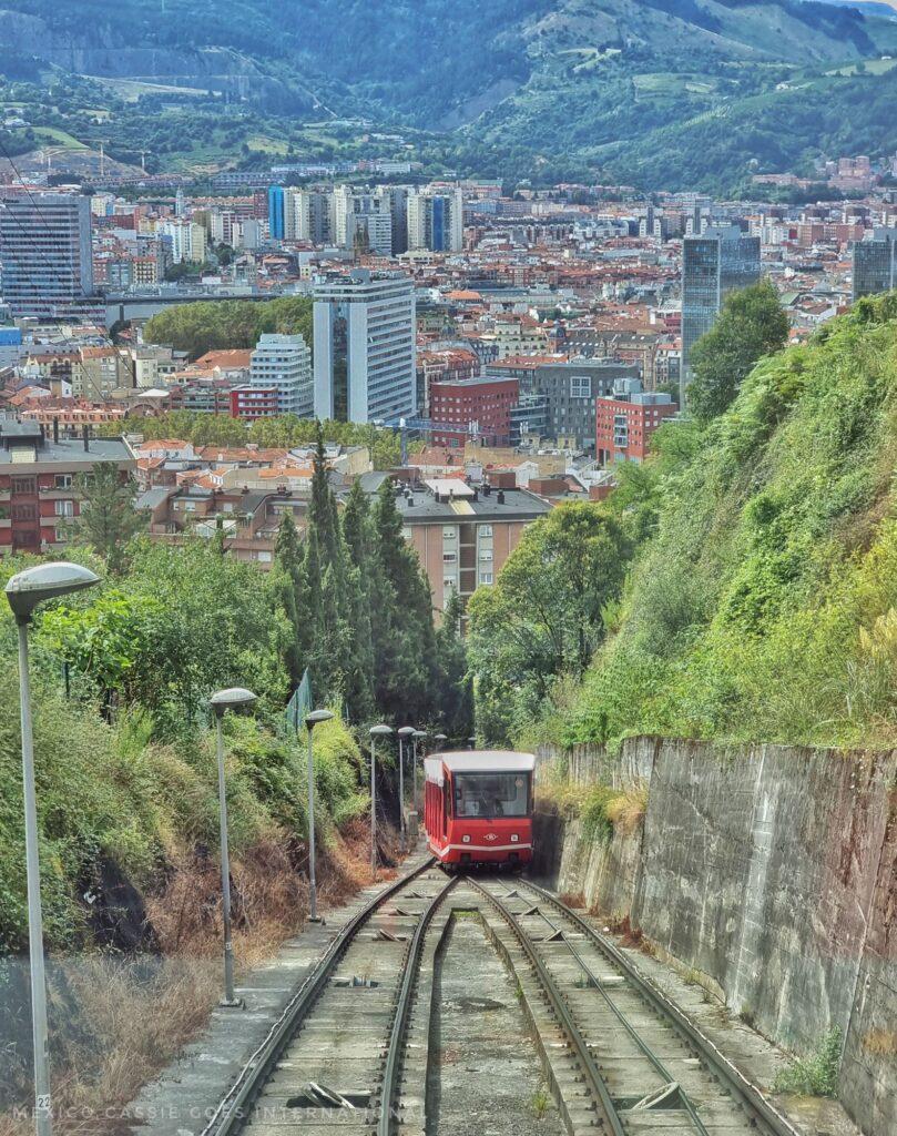 red funicular on tracks, greenery on either side and city below