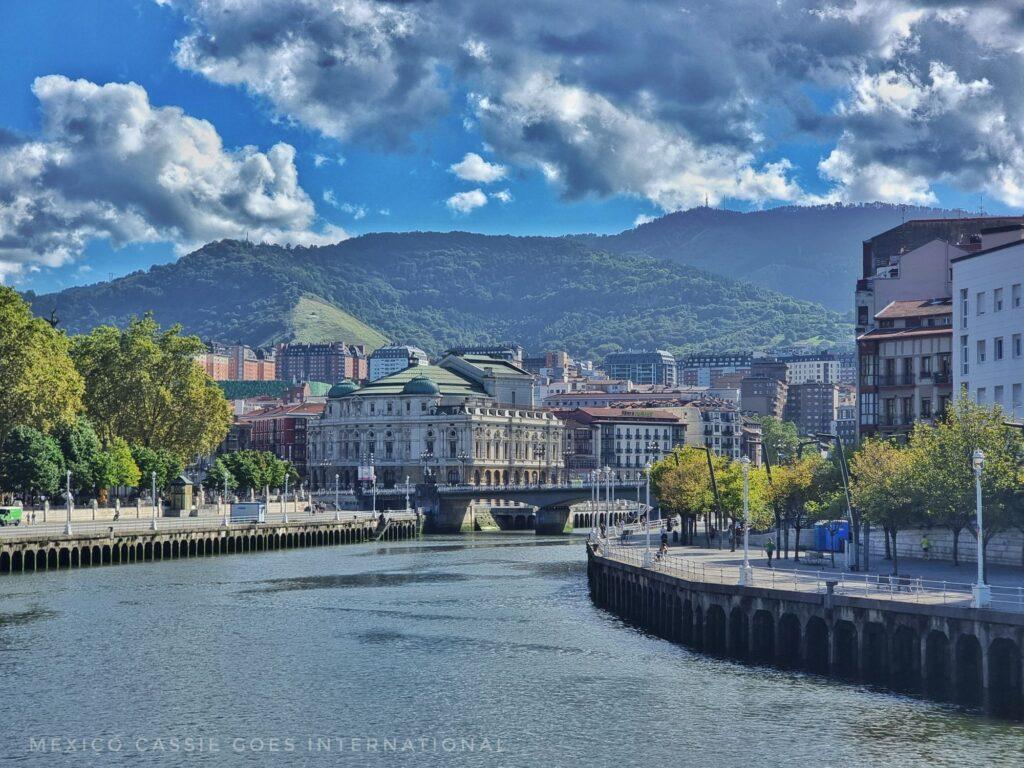 view of the river in Bilbao, green trees and buildings on banks, hills in distance
