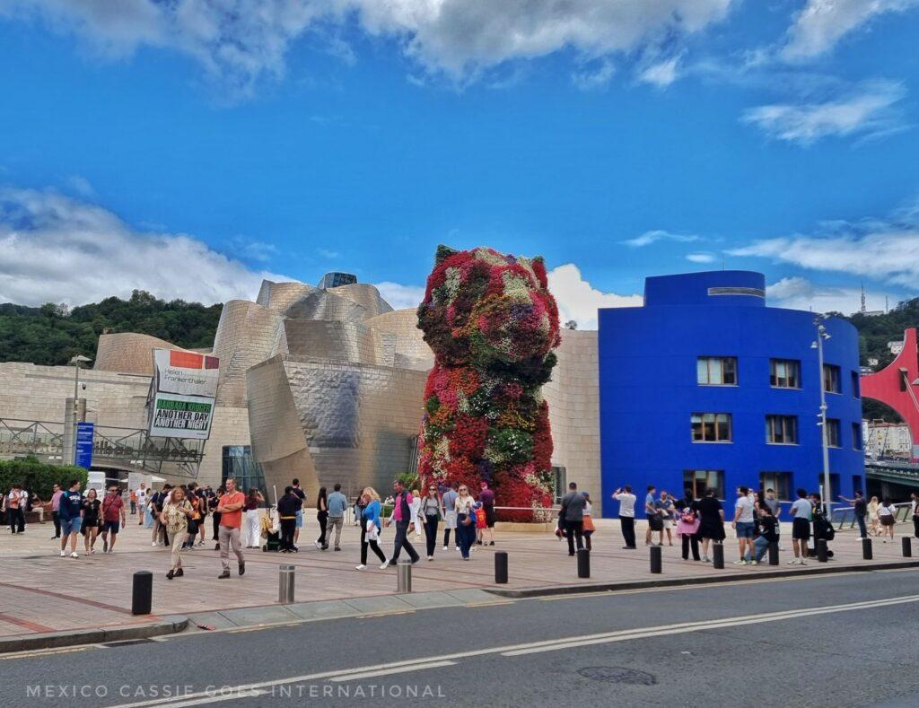 guggenheim museum from across the road. people milling around, flower puppy sculpture and blue building to right