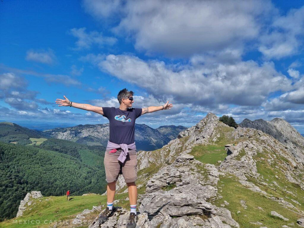 person in shorts and blue tshirt standing on rocks on top of mountain with blue sky and rolling green hills behind