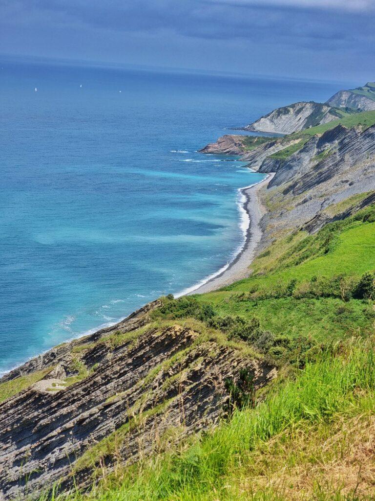 looking down from cliff top along a flysch coast in basque country - blue sea, striations of rock, green grass