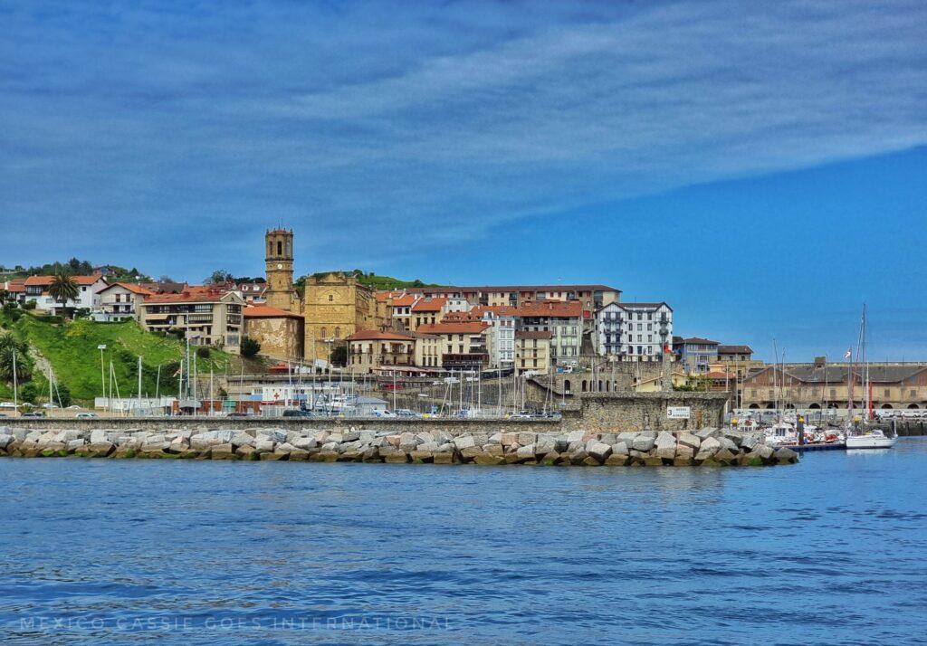 small town with harbour in front, photo taken from a boat on the calm sea - small boats, large castle, grassy area on left and buildings of small town in centre