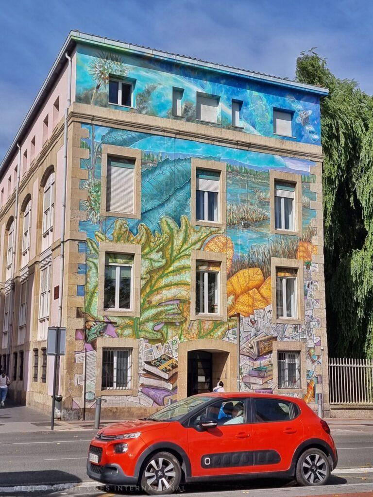 4 storey building with mural across it - books, nature designs of leaves and a river. red car parked in front