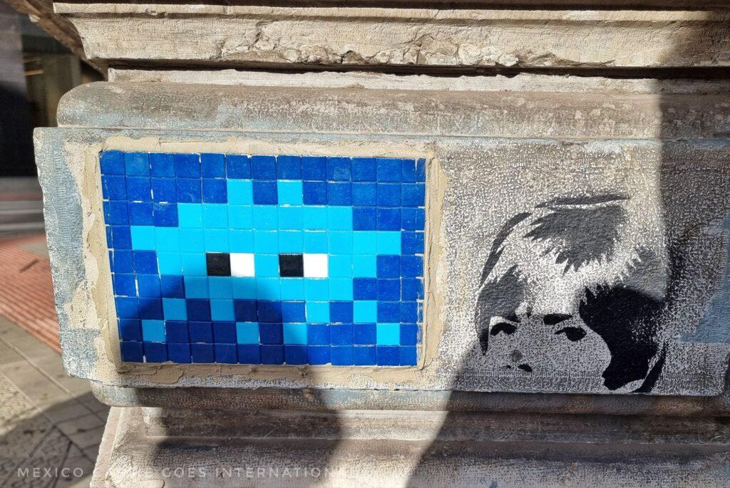 blue mosaic space invader on wall, next to a stencilled child's face