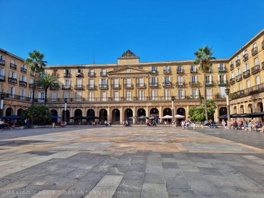 large and peaceful Spanish plaza. arches on ground floor, a few palm trees and scattered people under cafe umbrellas