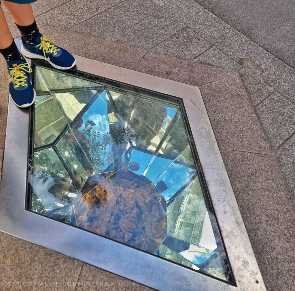 kid's feet and art installation in pavement - it's a ball of some sort in a glass 'cage' with silver metal frame
