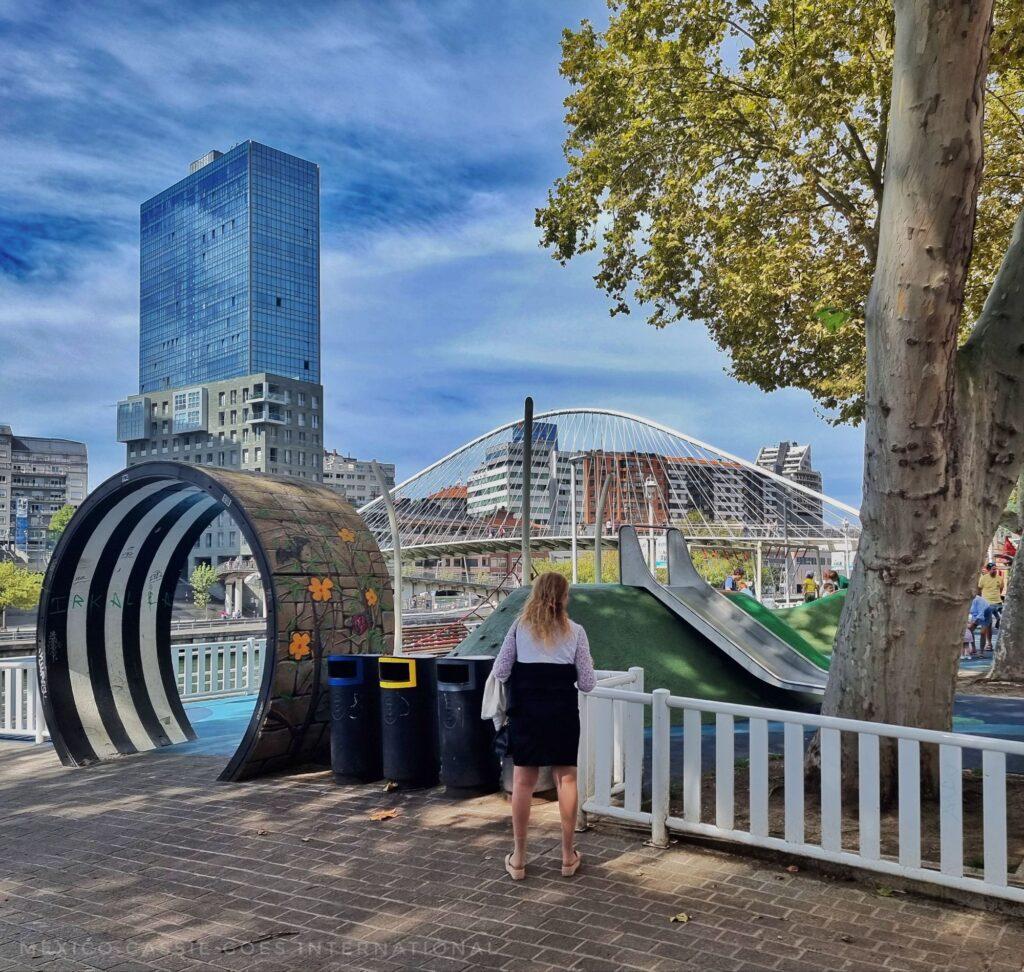 playground on riverside, woman standing by entrance (which is round) large tower on other side of river 